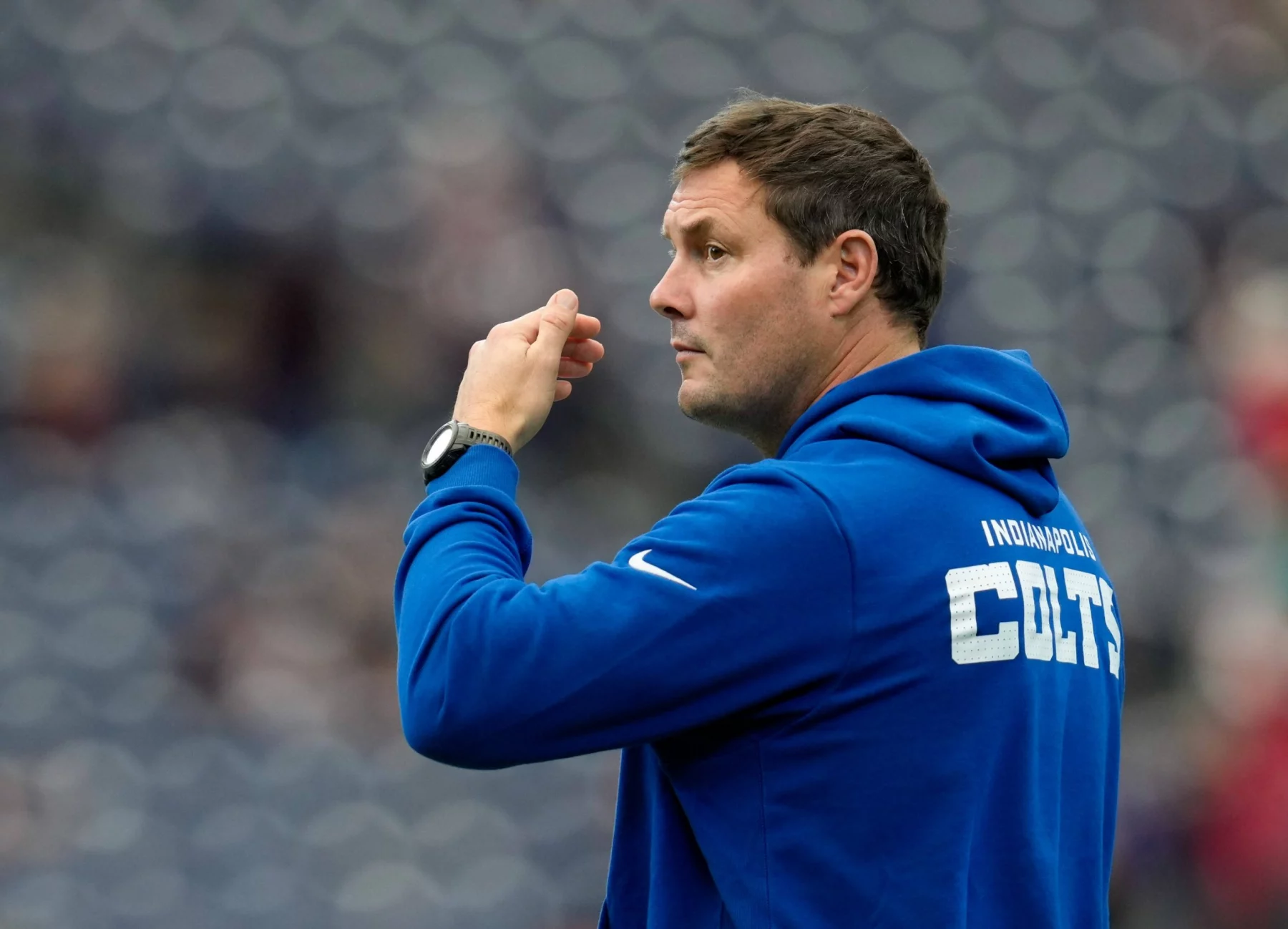 Philip Rivers - Ein Mann, der einen blauen "INDIANAPOLIS COLTS"-Kapuzenpulli trägt, steht im Profil und sieht aus wie Philip Rivers. Er scheint ein Sportereignis zu beobachten oder zu signalisieren, wobei die Sitze im Stadion im Hintergrund verschwimmen. Diese Beschreibung wurde mit der FootballR KI automatisch generiert.