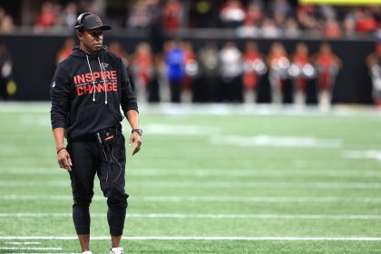 Falcons-Coach Raheem Morris steht in einem schwarzen "Inspire Change"-Kapuzenpulli, Kopfhörern, Mütze und Hose an der Seitenlinie. Er sieht konzentriert aus und hält ein Gerät in der Hand. Im verschwommenen Hintergrund sind Cheerleader und eine Stadionmenge zu sehen. Diese Beschreibung wurde mit der FootballR KI automatisch generiert.