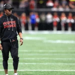 Falcons-Coach Raheem Morris steht in einem schwarzen "Inspire Change"-Kapuzenpulli, Kopfhörern, Mütze und Hose an der Seitenlinie. Er sieht konzentriert aus und hält ein Gerät in der Hand. Im verschwommenen Hintergrund sind Cheerleader und eine Stadionmenge zu sehen. Diese Beschreibung wurde mit der FootballR KI automatisch generiert.