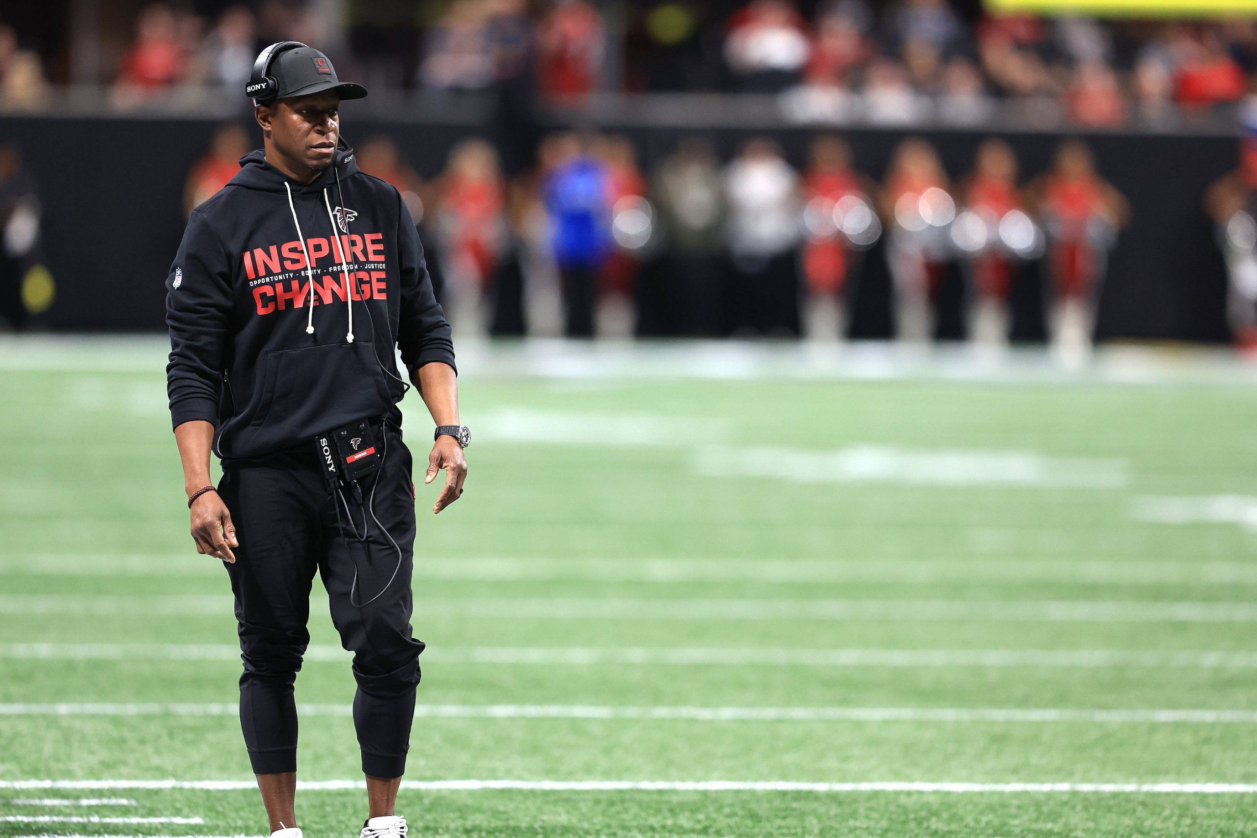 Falcons-Coach Raheem Morris steht in einem schwarzen "Inspire Change"-Kapuzenpulli, Kopfhörern, Mütze und Hose an der Seitenlinie. Er sieht konzentriert aus und hält ein Gerät in der Hand. Im verschwommenen Hintergrund sind Cheerleader und eine Stadionmenge zu sehen. Diese Beschreibung wurde mit der FootballR KI automatisch generiert.