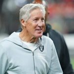 Pete Carroll - Ein älterer Mann mit grauem Haar lächelt, während er einen hellgrauen Raiders-Kapuzenpullover trägt. Er steht auf einem Football-Feld mit unscharfen Menschen und Lichtern im Hintergrund, was an eine Stadionkulisse erinnert, die an einen aufregenden Spieltag erinnert. Diese Beschreibung wurde mit der FootballR KI automatisch generiert.