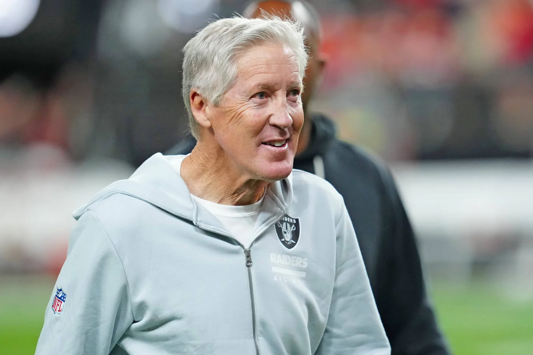 Pete Carroll - Ein älterer Mann mit grauem Haar lächelt, während er einen hellgrauen Raiders-Kapuzenpullover trägt. Er steht auf einem Football-Feld mit unscharfen Menschen und Lichtern im Hintergrund, was an eine Stadionkulisse erinnert, die an einen aufregenden Spieltag erinnert. Diese Beschreibung wurde mit der FootballR KI automatisch generiert.