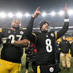 Zwei Footballspieler der Pittsburgh Steelers feiern auf dem Spielfeld in ihren schwarz-gelben Trikots. Der eine streckt den Arm aus, der andere zeigt nach oben und zeigt seinen Stolz auf die AFC North, während im Hintergrund die Sitze und Lichter des Stadions leuchten. Diese Beschreibung wurde mit der FootballR KI automatisch generiert.