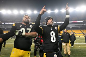 Zwei Footballspieler der Pittsburgh Steelers feiern auf dem Spielfeld in ihren schwarz-gelben Trikots. Der eine streckt den Arm aus, der andere zeigt nach oben und zeigt seinen Stolz auf die AFC North, während im Hintergrund die Sitze und Lichter des Stadions leuchten. Diese Beschreibung wurde mit der FootballR KI automatisch generiert.