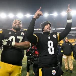 Zwei Footballspieler der Pittsburgh Steelers feiern auf dem Spielfeld in ihren schwarz-gelben Trikots. Der eine streckt den Arm aus, der andere zeigt nach oben und zeigt seinen Stolz auf die AFC North, während im Hintergrund die Sitze und Lichter des Stadions leuchten. Diese Beschreibung wurde mit der FootballR KI automatisch generiert.