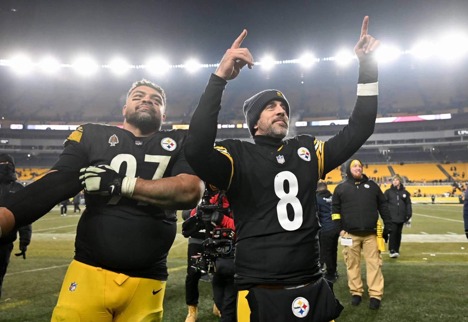 Zwei Footballspieler der Pittsburgh Steelers feiern auf dem Spielfeld in ihren schwarz-gelben Trikots. Der eine streckt den Arm aus, der andere zeigt nach oben und zeigt seinen Stolz auf die AFC North, während im Hintergrund die Sitze und Lichter des Stadions leuchten. Diese Beschreibung wurde mit der FootballR KI automatisch generiert.