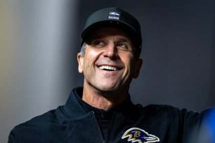 Ein lächelnder Mann mit schwarzer Baltimore-Ravens-Mütze und -Jacke steht im Freien vor einem unscharfen Hintergrund. Auf seiner Jacke ist das Ravens-Logo zu sehen, das an die langjährige Verpflichtung von John Harbaugh gegenüber dem Team erinnert. Diese Beschreibung wurde mit der FootballR KI automatisch generiert.