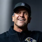 Ein lächelnder Mann mit schwarzer Baltimore-Ravens-Mütze und -Jacke steht im Freien vor einem unscharfen Hintergrund. Auf seiner Jacke ist das Ravens-Logo zu sehen, das an die langjährige Verpflichtung von John Harbaugh gegenüber dem Team erinnert. Diese Beschreibung wurde mit der FootballR KI automatisch generiert.