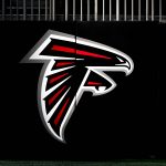 Ein großes Atlanta Falcons-Logo mit einem stilisierten schwarzen, roten und weißen Falken im Flug ist auf einer dunklen Wand zu sehen. Am oberen Rand des Bildes über dem Logo sind vertikale Metallstäbe zu sehen, die Ian Cunningham Verbindung zum Team hervorheben. Diese Beschreibung wurde mit der FootballR KI automatisch generiert.