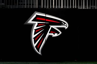 Ein großes Atlanta Falcons-Logo mit einem stilisierten schwarzen, roten und weißen Falken im Flug ist auf einer dunklen Wand zu sehen. Am oberen Rand des Bildes über dem Logo sind vertikale Metallstäbe zu sehen, die Ian Cunningham Verbindung zum Team hervorheben. Diese Beschreibung wurde mit der FootballR KI automatisch generiert.
