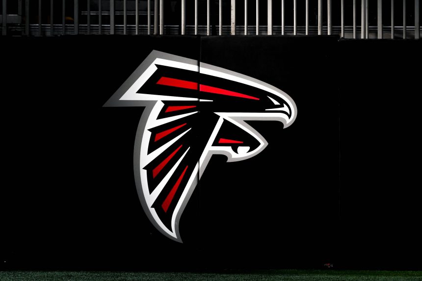 Ein großes Atlanta Falcons-Logo mit einem stilisierten schwarzen, roten und weißen Falken im Flug ist auf einer dunklen Wand zu sehen. Am oberen Rand des Bildes über dem Logo sind vertikale Metallstäbe zu sehen, die Ian Cunningham Verbindung zum Team hervorheben. Diese Beschreibung wurde mit der FootballR KI automatisch generiert.