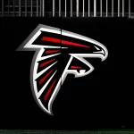 Ein großes Atlanta Falcons-Logo mit einem stilisierten schwarzen, roten und weißen Falken im Flug ist auf einer dunklen Wand zu sehen. Am oberen Rand des Bildes über dem Logo sind vertikale Metallstäbe zu sehen, die Ian Cunningham Verbindung zum Team hervorheben. Diese Beschreibung wurde mit der FootballR KI automatisch generiert.