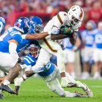 Ein Footballspieler der Miami Hurricanes in einer weißen Uniform wird während eines College-Football-Spiels von zwei Spielern der Ole Miss in blauen Uniformen angegriffen. Der Miami-Spieler hält den Ball fest umklammert, als er zu Boden gebracht wird. Im unscharfen Hintergrund sind die Fans zu sehen. Diese Beschreibung wurde mit der FootballR KI automatisch generiert.