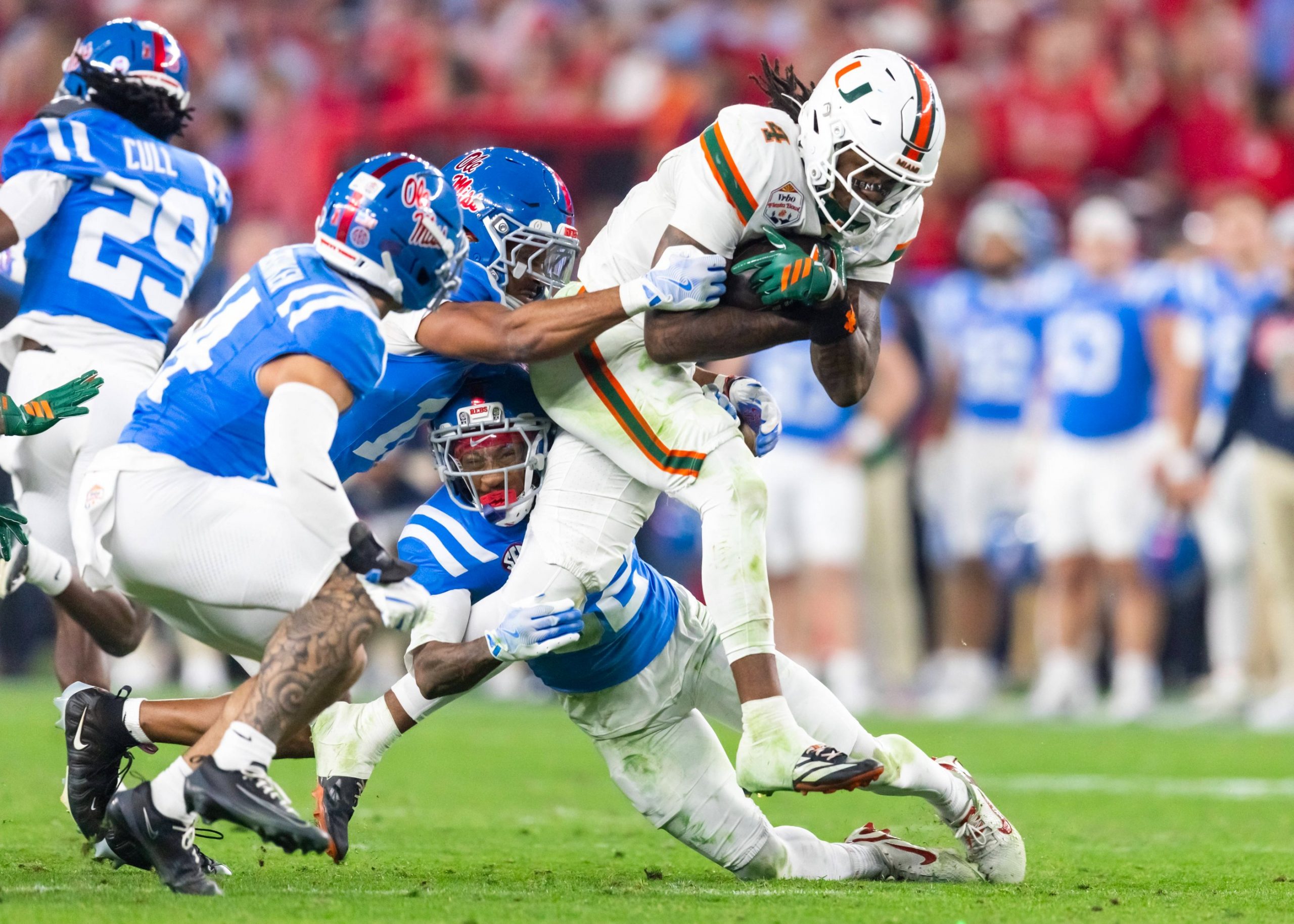 Ein Footballspieler der Miami Hurricanes in einer weißen Uniform wird während eines College-Football-Spiels von zwei Spielern der Ole Miss in blauen Uniformen angegriffen. Der Miami-Spieler hält den Ball fest umklammert, als er zu Boden gebracht wird. Im unscharfen Hintergrund sind die Fans zu sehen. Diese Beschreibung wurde mit der FootballR KI automatisch generiert.