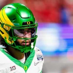 Oregon-Quarterback Dante Moore, der einen grün-gelben Helm mit einem "O"-Logo und einem Goodyear Cotton Bowl-Aufnäher trägt, hebt sich vor einem verschwommenen rot-lila Hintergrund ab - ein potenzieller Topkandidat für den NFL-Draft 2026. Diese Beschreibung wurde mit der FootballR KI automatisch generiert.
