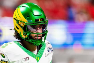 Oregon-Quarterback Dante Moore, der einen grün-gelben Helm mit einem "O"-Logo und einem Goodyear Cotton Bowl-Aufnäher trägt, hebt sich vor einem verschwommenen rot-lila Hintergrund ab - ein potenzieller Topkandidat für den NFL-Draft 2026. Diese Beschreibung wurde mit der FootballR KI automatisch generiert.
