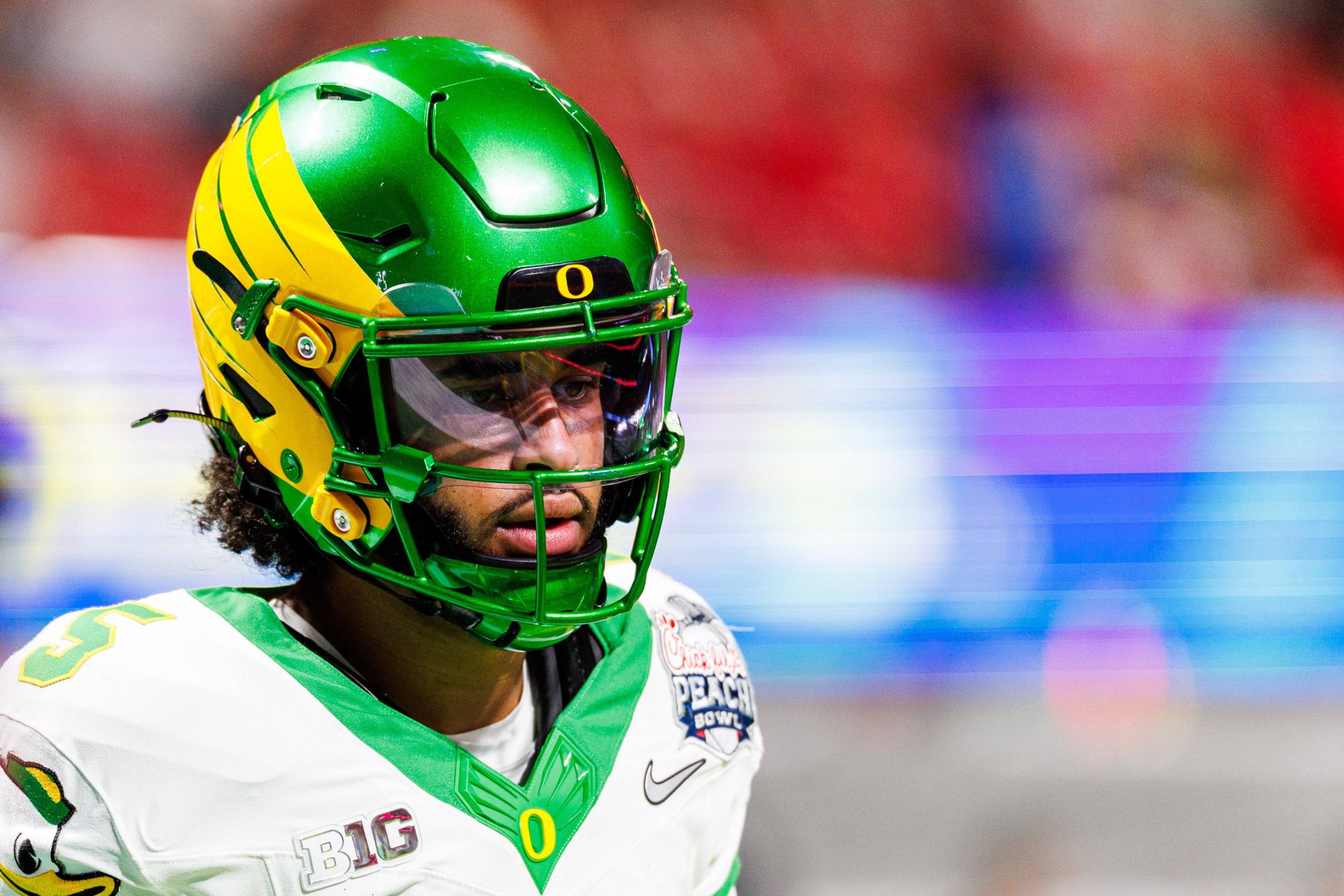 Oregon-Quarterback Dante Moore, der einen grün-gelben Helm mit einem "O"-Logo und einem Goodyear Cotton Bowl-Aufnäher trägt, hebt sich vor einem verschwommenen rot-lila Hintergrund ab - ein potenzieller Topkandidat für den NFL-Draft 2026. Diese Beschreibung wurde mit der FootballR KI automatisch generiert.