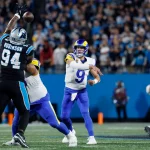Rams-Quarterback Matthew Stafford in einer weiß-blauen Uniform wirft einen Football, während ein Panthers-Verteidiger in Schwarz zum Block springt. Im Hintergrund sind andere Spieler und eine Menge von Fans in einem blau beleuchteten Stadion zu sehen. Diese Beschreibung wurde mit der FootballR KI automatisch generiert.