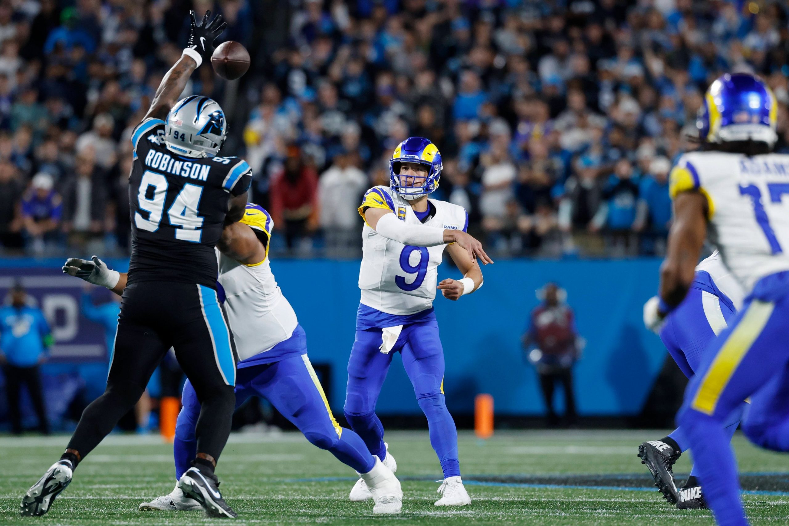 Rams-Quarterback Matthew Stafford in einer weiß-blauen Uniform wirft einen Football, während ein Panthers-Verteidiger in Schwarz zum Block springt. Im Hintergrund sind andere Spieler und eine Menge von Fans in einem blau beleuchteten Stadion zu sehen. Diese Beschreibung wurde mit der FootballR KI automatisch generiert.