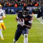 Ein Spieler der Chicago Bears rennt mit einem Football durch die Gegend, während ein Verteidiger der Green Bay Packers ihn während eines hitzigen Divisional-Playoff-Spiels verfolgt. Zuschauer und Schiedsrichter beobachten das Spiel vom Spielfeldrand aus unter hellen Lichtern; das Spiel findet bei Nacht statt. Diese Beschreibung wurde mit der FootballR KI automatisch generiert.
