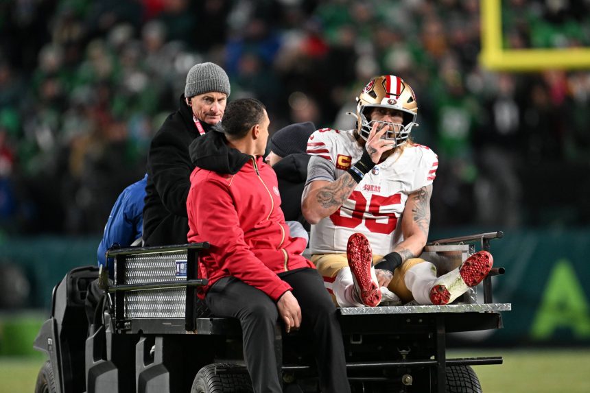 Der Spieler der San Francisco 49ers, George Kittle, in voller Montur und mit Helm, sitzt nach einem möglichen Achillessehnenriss auf einem Krankenwagen und legt sein rechtes Bein hoch. Zwei Mitarbeiter helfen ihm, während die verschwommene Menge und die Stadionbeleuchtung den Hintergrund ausfüllen. Diese Beschreibung wurde mit der FootballR KI automatisch generiert.