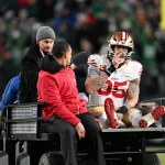 Der Spieler der San Francisco 49ers, George Kittle, in voller Montur und mit Helm, sitzt nach einem möglichen Achillessehnenriss auf einem Krankenwagen und legt sein rechtes Bein hoch. Zwei Mitarbeiter helfen ihm, während die verschwommene Menge und die Stadionbeleuchtung den Hintergrund ausfüllen. Diese Beschreibung wurde mit der FootballR KI automatisch generiert.