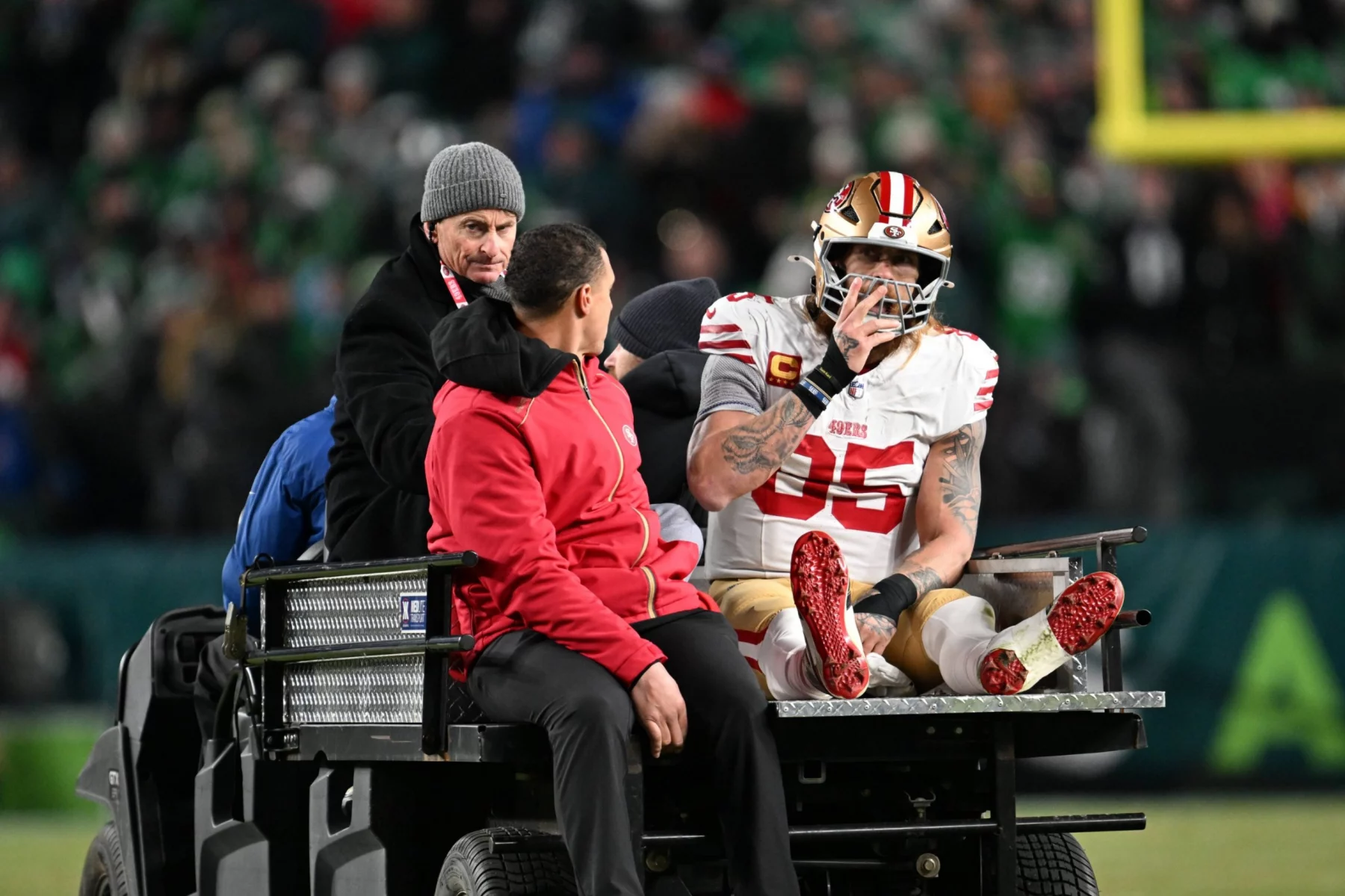 Der Spieler der San Francisco 49ers, George Kittle, in voller Montur und mit Helm, sitzt nach einem möglichen Achillessehnenriss auf einem Krankenwagen und legt sein rechtes Bein hoch. Zwei Mitarbeiter helfen ihm, während die verschwommene Menge und die Stadionbeleuchtung den Hintergrund ausfüllen. Diese Beschreibung wurde mit der FootballR KI automatisch generiert.