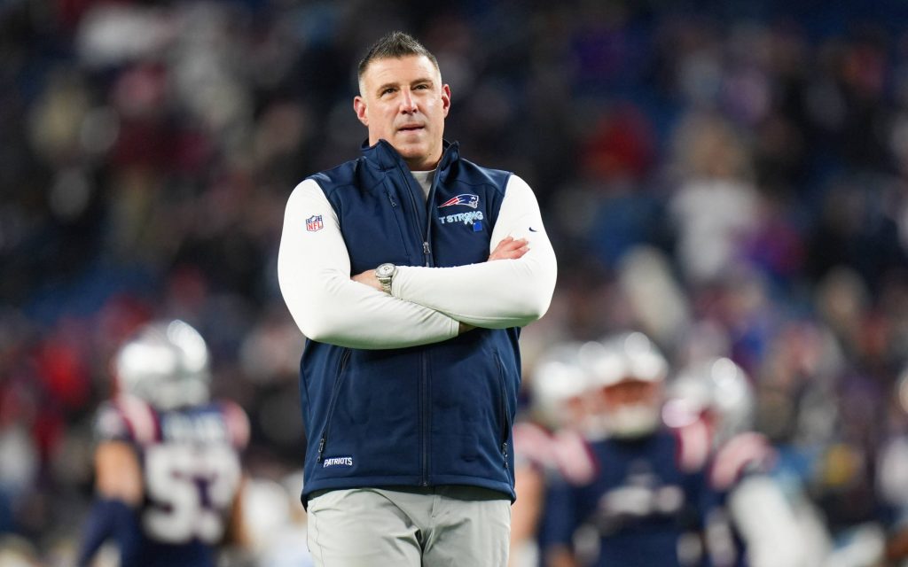 Mike Vrabel - Wild Card - Ein Mann steht mit verschränkten Armen auf einem Football-Feld und trägt ein weißes langärmeliges Hemd unter einer blauen Weste der New England Patriots während der NFL Playoffs. Im unscharfen Hintergrund sind die Spieler der Patriots und die mit Fans gefüllten Stadionplätze zu sehen. Diese Beschreibung wurde mit der FootballR KI automatisch generiert.