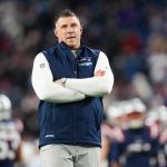 Mike Vrabel - Wild Card - Ein Mann steht mit verschränkten Armen auf einem Football-Feld und trägt ein weißes langärmeliges Hemd unter einer blauen Weste der New England Patriots während der NFL Playoffs. Im unscharfen Hintergrund sind die Spieler der Patriots und die mit Fans gefüllten Stadionplätze zu sehen. Diese Beschreibung wurde mit der FootballR KI automatisch generiert.