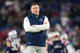 Mike Vrabel - Wild Card - Ein Mann steht mit verschränkten Armen auf einem Football-Feld und trägt ein weißes langärmeliges Hemd unter einer blauen Weste der New England Patriots während der NFL Playoffs. Im unscharfen Hintergrund sind die Spieler der Patriots und die mit Fans gefüllten Stadionplätze zu sehen. Diese Beschreibung wurde mit der FootballR KI automatisch generiert.