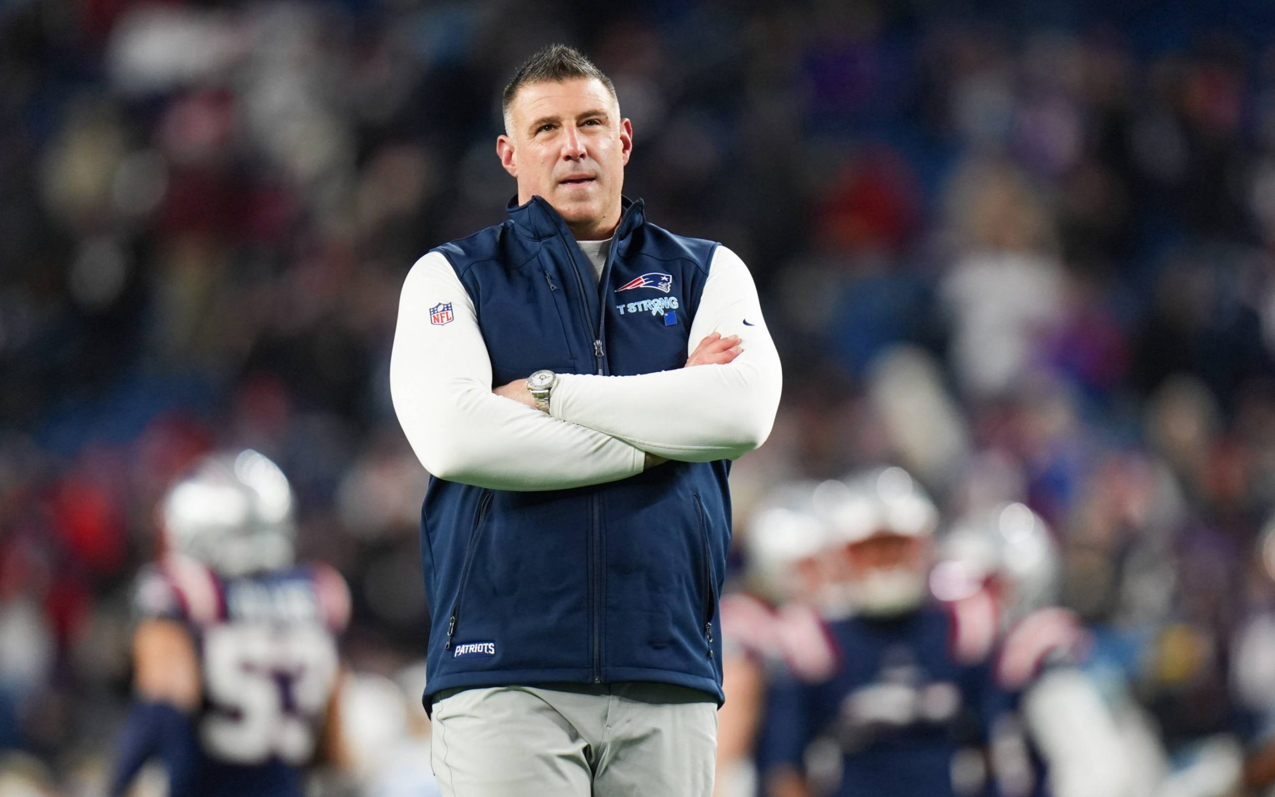 Mike Vrabel - Wild Card - Ein Mann steht mit verschränkten Armen auf einem Football-Feld und trägt ein weißes langärmeliges Hemd unter einer blauen Weste der New England Patriots während der NFL Playoffs. Im unscharfen Hintergrund sind die Spieler der Patriots und die mit Fans gefüllten Stadionplätze zu sehen. Diese Beschreibung wurde mit der FootballR KI automatisch generiert.