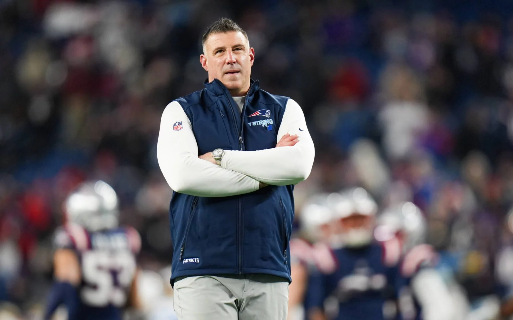 Mike Vrabel - Wild Card - Ein Mann steht mit verschränkten Armen auf einem Football-Feld und trägt ein weißes langärmeliges Hemd unter einer blauen Weste der New England Patriots während der NFL Playoffs. Im unscharfen Hintergrund sind die Spieler der Patriots und die mit Fans gefüllten Stadionplätze zu sehen. Diese Beschreibung wurde mit der FootballR KI automatisch generiert.