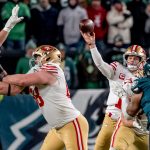 Brock Purdy - Ein Quarterback der San Francisco 49ers wirft einen Football, während die Verteidiger der Eagles auf ihn zustürmen. Ein Lineman der 49ers blockt einen Spieler der Eagles. Die unscharfe grüne Menschenmenge im Hintergrund unterstreicht diesen intensiven Spielmoment, trotz des Risikos von Verletzungen. Diese Beschreibung wurde mit der FootballR KI automatisch generiert.