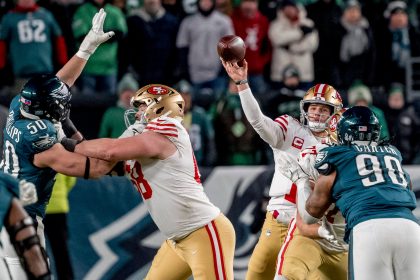 Brock Purdy - Ein Quarterback der San Francisco 49ers wirft einen Football, während die Verteidiger der Eagles auf ihn zustürmen. Ein Lineman der 49ers blockt einen Spieler der Eagles. Die unscharfe grüne Menschenmenge im Hintergrund unterstreicht diesen intensiven Spielmoment, trotz des Risikos von Verletzungen. Diese Beschreibung wurde mit der FootballR KI automatisch generiert.