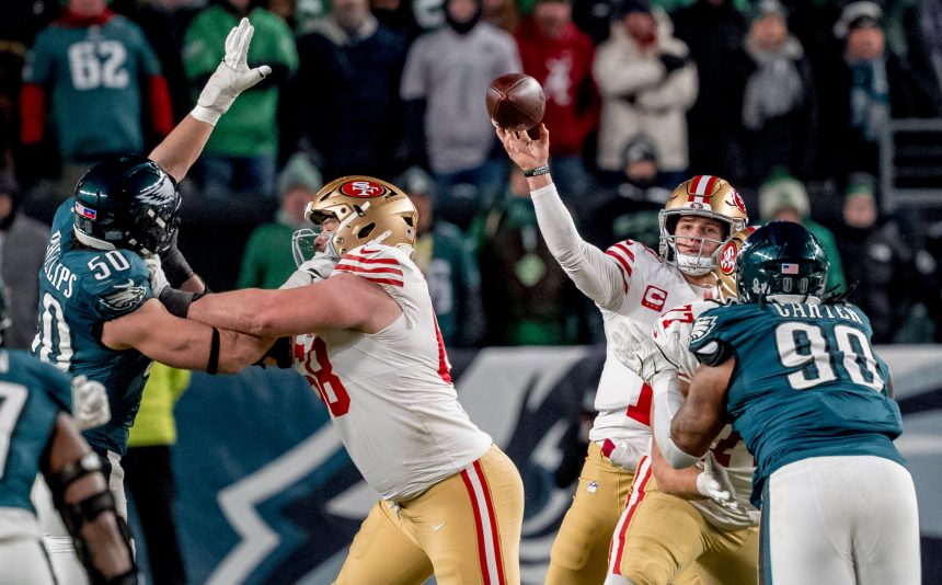 Brock Purdy - Ein Quarterback der San Francisco 49ers wirft einen Football, während die Verteidiger der Eagles auf ihn zustürmen. Ein Lineman der 49ers blockt einen Spieler der Eagles. Die unscharfe grüne Menschenmenge im Hintergrund unterstreicht diesen intensiven Spielmoment, trotz des Risikos von Verletzungen. Diese Beschreibung wurde mit der FootballR KI automatisch generiert.