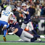 Drake Maye - Ein Quarterback der New England Patriots in einer Marineuniform wirft einen Football, während ein Teamkollege auf den Rasen fällt und einen Verteidiger der Los Angeles Chargers blockt. Das Spiel findet auf einem hell erleuchteten Football-Feld statt, im Hintergrund die Patriots-Fans. Diese Beschreibung wurde mit der FootballR KI automatisch generiert.