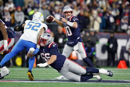Drake Maye - Ein Quarterback der New England Patriots in einer Marineuniform wirft einen Football, während ein Teamkollege auf den Rasen fällt und einen Verteidiger der Los Angeles Chargers blockt. Das Spiel findet auf einem hell erleuchteten Football-Feld statt, im Hintergrund die Patriots-Fans. Diese Beschreibung wurde mit der FootballR KI automatisch generiert.
