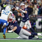 Drake Maye - Ein Quarterback der New England Patriots in einer Marineuniform wirft einen Football, während ein Teamkollege auf den Rasen fällt und einen Verteidiger der Los Angeles Chargers blockt. Das Spiel findet auf einem hell erleuchteten Football-Feld statt, im Hintergrund die Patriots-Fans. Diese Beschreibung wurde mit der FootballR KI automatisch generiert.