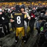 Houston Texans - Zwei American-Football-Spieler umarmen sich nach einem Spiel der Wild Card Round auf dem Spielfeld. Der Spieler, der der Kamera zugewandt ist, trägt ein schwarz-gelbes Steelers-Trikot mit der Aufschrift "RODGERS 8" auf dem Rücken, umgeben von Fotografen, Reportern und jubelnden Fans auf den Sitzen im Stadion. Diese Beschreibung wurde mit der FootballR KI automatisch generiert.