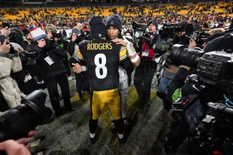 Houston Texans - Aaron Rodgers - Zwei American-Football-Spieler umarmen sich nach einem Spiel der Wild Card Round auf dem Spielfeld. Der Spieler, der der Kamera zugewandt ist, trägt ein schwarz-gelbes Steelers-Trikot mit der Aufschrift "RODGERS 8" auf dem Rücken, umgeben von Fotografen, Reportern und jubelnden Fans auf den Sitzen im Stadion. Diese Beschreibung wurde mit der FootballR KI automatisch generiert.