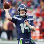 Sam Darnold - Ein Footballspieler im Trikot der Seattle Seahawks mit der Nummer 14 befindet sich mitten im Wurf während eines Spiels. Mit der Energie eines Playoff-Sieges trägt er einen dunklen Helm und hält den Football in der rechten Hand, während rot gekleidete Spieler im Hintergrund verschwimmen. Diese Beschreibung wurde mit der FootballR KI automatisch generiert.