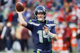 Sam Darnold - Ein Footballspieler im Trikot der Seattle Seahawks mit der Nummer 14 befindet sich mitten im Wurf während eines Spiels. Mit der Energie eines Playoff-Sieges trägt er einen dunklen Helm und hält den Football in der rechten Hand, während rot gekleidete Spieler im Hintergrund verschwimmen. Diese Beschreibung wurde mit der FootballR KI automatisch generiert.