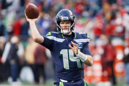 Sam Darnold - Ein Footballspieler im Trikot der Seattle Seahawks mit der Nummer 14 befindet sich mitten im Wurf während eines Spiels. Mit der Energie eines Playoff-Sieges trägt er einen dunklen Helm und hält den Football in der rechten Hand, während rot gekleidete Spieler im Hintergrund verschwimmen. Diese Beschreibung wurde mit der FootballR KI automatisch generiert.