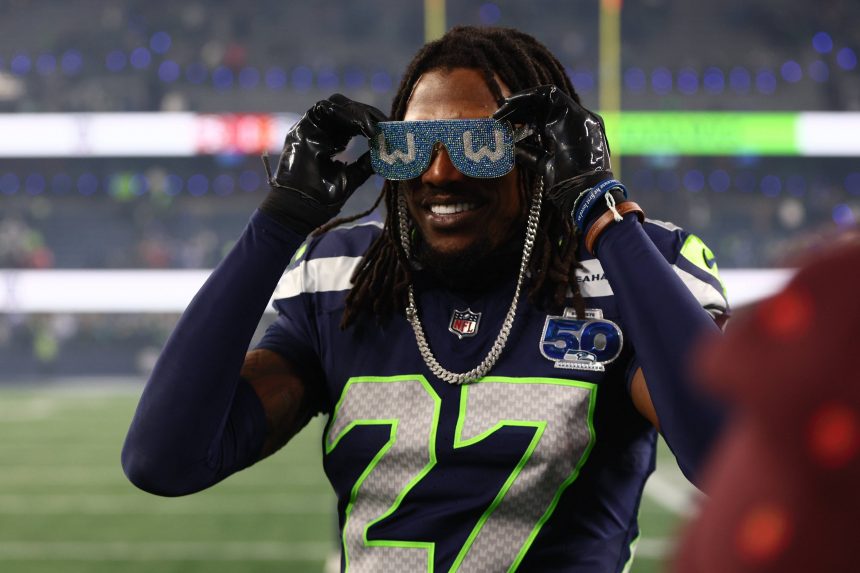 Ein lächelnder Footballspieler der Seattle Seahawks, Riq Woolen, mit der Nummer 27 und einem Aufnäher der 50. Saison hält sich eine große blaue Glitzer-"W"-Brille vors Gesicht. Handschuhe, eine Halskette und lange geflochtene Haare vervollständigen sein Aussehen vor der unscharfen Stadionkulisse. Diese Beschreibung wurde mit der FootballR KI automatisch generiert.