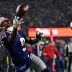 Ein Football-Spieler in einer marineblauen New England Patriots Uniform, möglicherweise Drake Maye, streckt die Hand aus, um einen Ball während eines verschneiten Nachtspiels zu fangen, während ein Texans-Verteidiger in Weiß zusieht. Die Szene ist intensiv, mit Schneefall und verschwommenen Zuschauern im Hintergrund. Diese Beschreibung wurde mit der FootballR KI automatisch generiert.