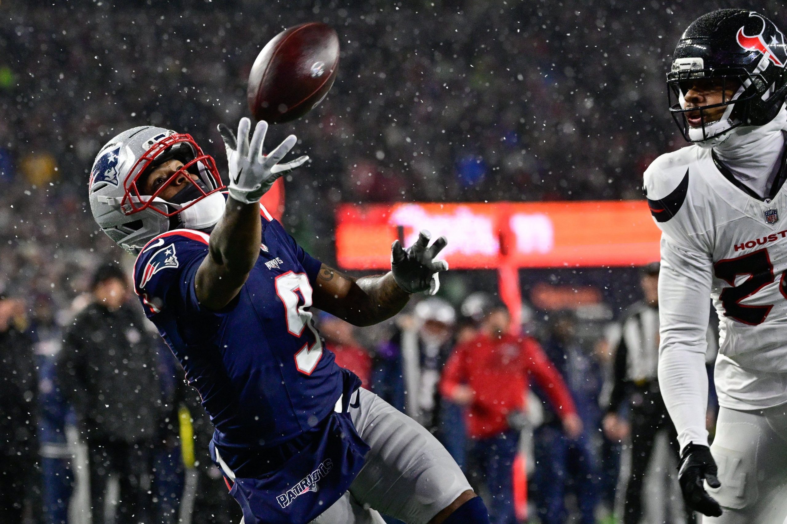 Ein Football-Spieler in einer marineblauen New England Patriots Uniform, möglicherweise Drake Maye, streckt die Hand aus, um einen Ball während eines verschneiten Nachtspiels zu fangen, während ein Texans-Verteidiger in Weiß zusieht. Die Szene ist intensiv, mit Schneefall und verschwommenen Zuschauern im Hintergrund. Diese Beschreibung wurde mit der FootballR KI automatisch generiert.