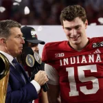 Ein lächelnder Indiana Footballspieler, Fernando Mendoza, mit der Trikotnummer 15 steht neben einem Interviewer, der ein Mikrofon hält. Konfetti fällt, während sie neben einer goldenen Meisterschaftstrophäe den nationalen Titel feiern. Der Spieler trägt einen Aufnäher mit der Aufschrift "2026"; das Stadion hinter ihnen ist unscharf. Diese Beschreibung wurde mit der FootballR KI automatisch generiert.