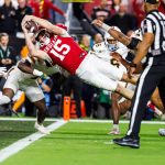 Indiana QB Fernando Mendoza (Nr. 15) springt waagerecht über die Torlinie und streckt den Ball mit der rechten Hand aus. Die Verteidiger in Weiß verfolgen ihn, während ein Offizieller zuschaut. Die Fans sehen ihren potenziellen Star des NFL Draft 2026 in Aktion. Diese Beschreibung wurde mit der FootballR KI automatisch generiert.