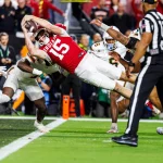 Indiana QB Fernando Mendoza (Nr. 15) springt waagerecht über die Torlinie und streckt den Ball mit der rechten Hand aus. Die Verteidiger in Weiß verfolgen ihn, während ein Offizieller zuschaut. Die Fans sehen ihren potenziellen Star des NFL Draft 2026 in Aktion. Diese Beschreibung wurde mit der FootballR KI automatisch generiert.