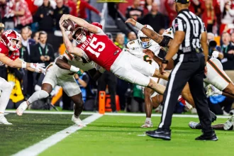 Indiana QB Fernando Mendoza (Nr. 15) springt waagerecht über die Torlinie und streckt den Ball mit der rechten Hand aus. Die Verteidiger in Weiß verfolgen ihn, während ein Offizieller zuschaut. Die Fans sehen ihren potenziellen Star des NFL Draft 2026 in Aktion. Diese Beschreibung wurde mit der FootballR KI automatisch generiert.